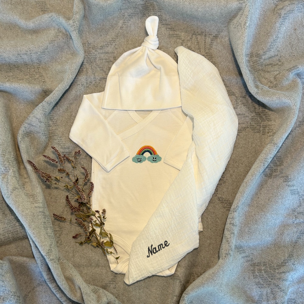 Ein besticktes Geschenkset für Babys in der Farbe Weiß, das ein Bio-Baumwolle Baby-Body, ein personalisiertes Bio-Musselin-Tuch mit unbehandeltem Holzring, eine Bio-Baumwolle Mütze und eine Kontrastkarte für ein Baby beinhaltet.